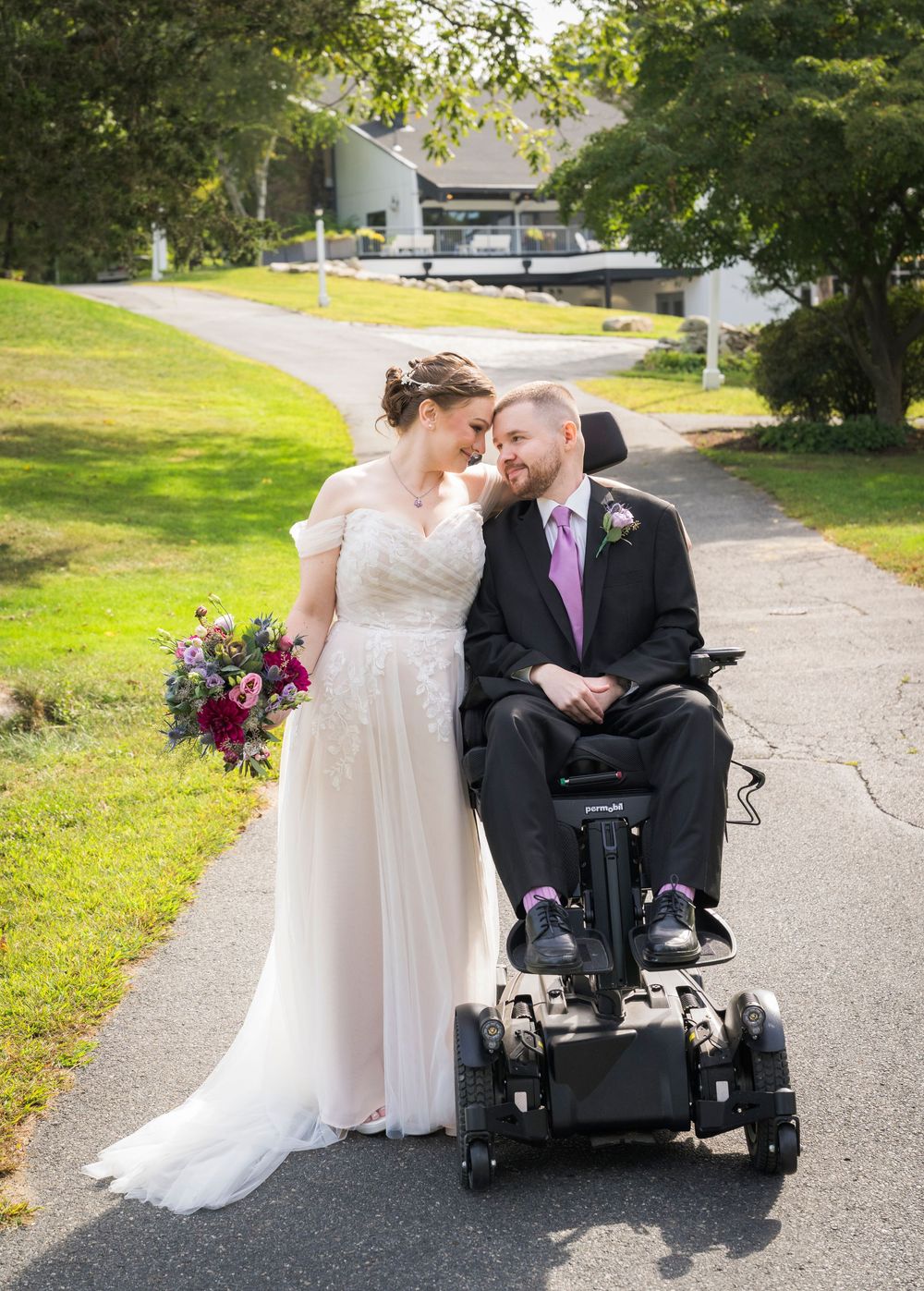 Bride in white lace gown holds colorful bouquet beside groom in wheelchair on tree-lined pathway.