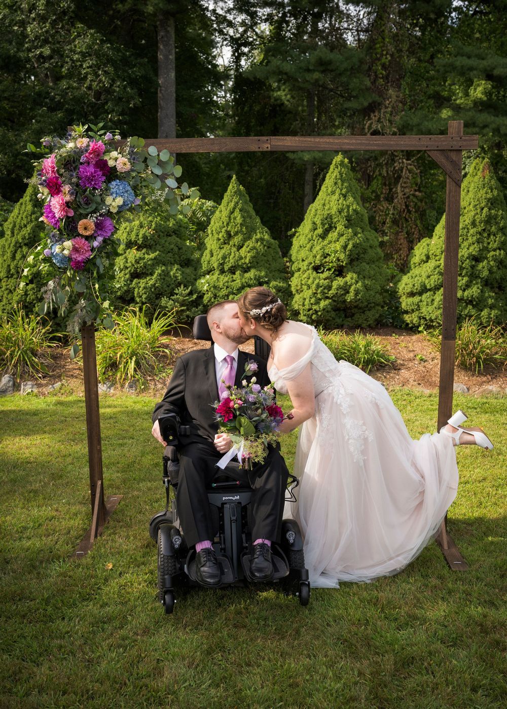 Bride in white lace gown kisses groom in wheelchair under floral arch with colorful blooms at outdoor wedding.