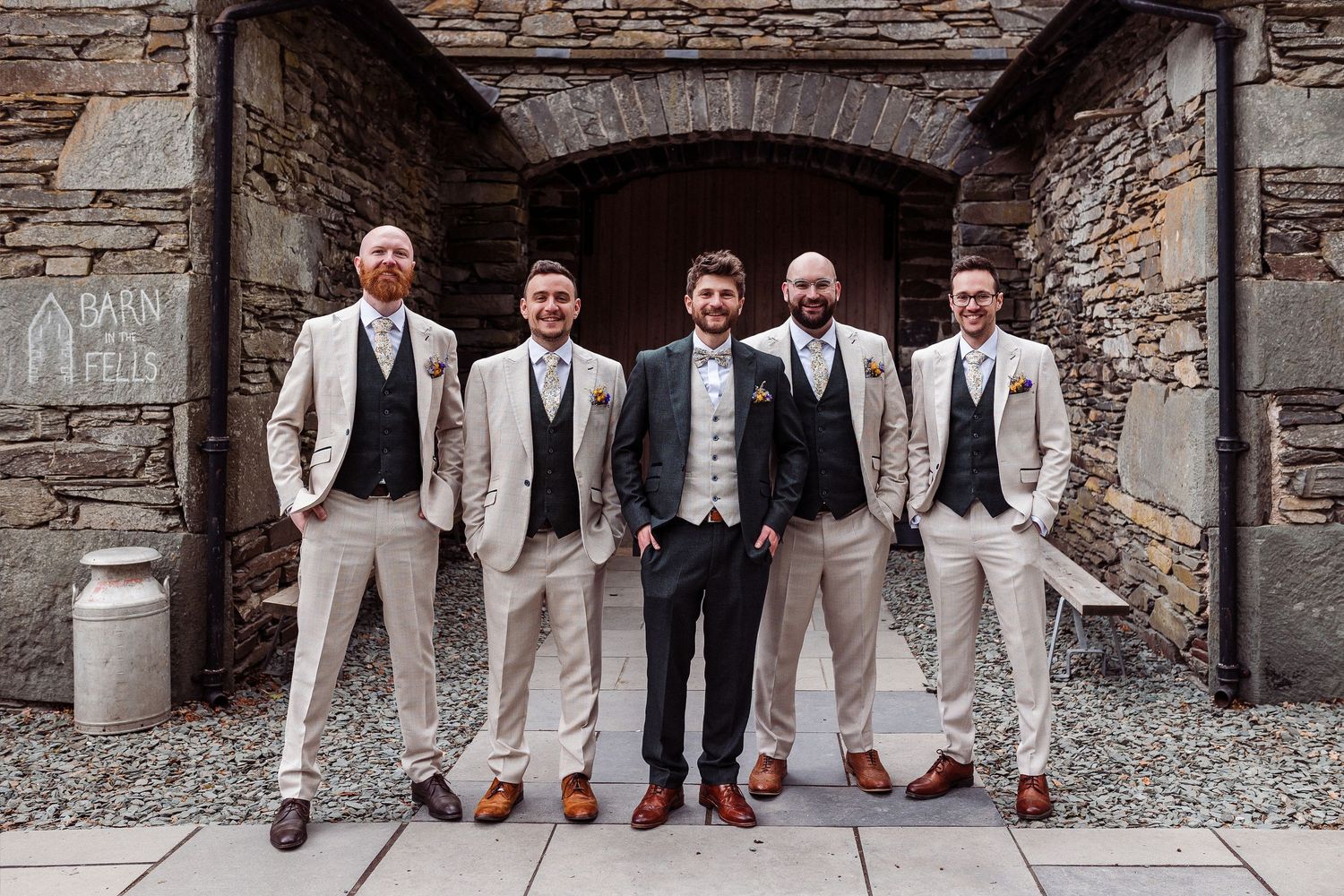 Wedding party poses before rustic wooden doors, bride and groom centered, attendants in gold, orange, and purple gowns with tan suits.