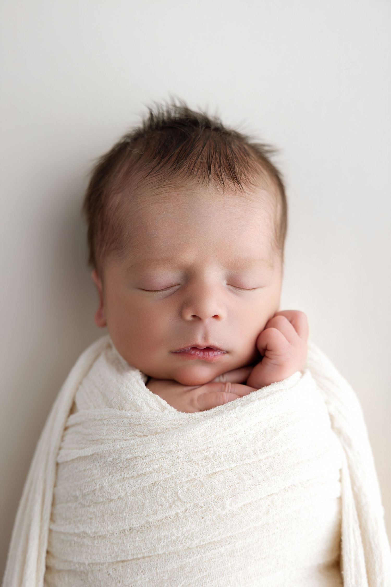 Peaceful newborn baby swaddled in a soft white knit wrap, sleeping with tiny hand tucked under chin.