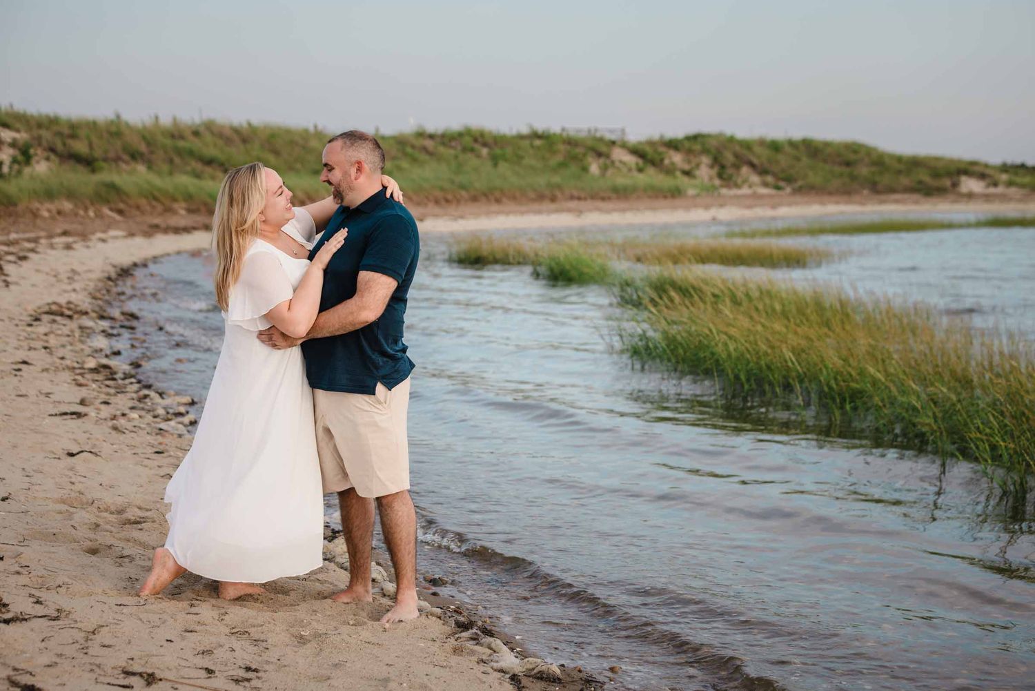 Engaged couple embracing at the water's edge at Duxbury Beach, Massachusetts, with marsh grass and tidal creek behind them.