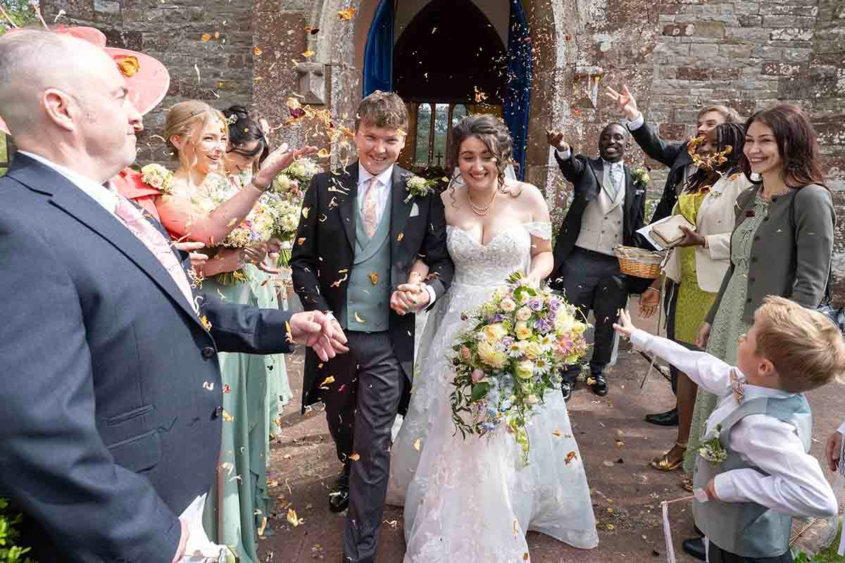 Bride and groom walk through confetti shower outside stone church surrounded by smiling wedding guests.