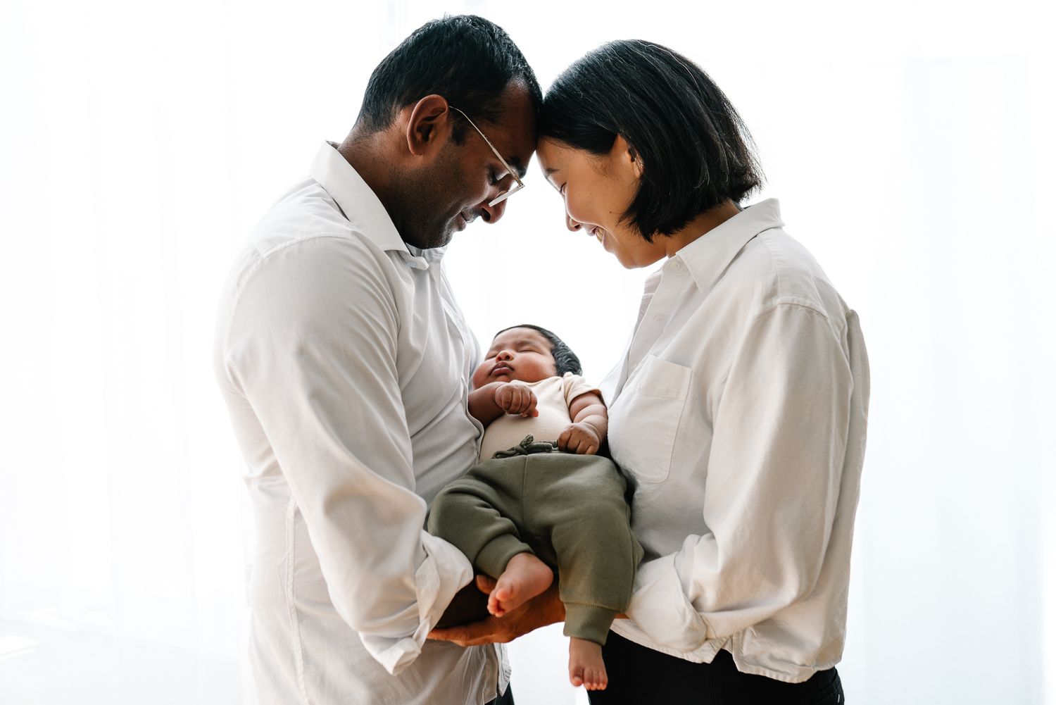 Interracial couple lovingly holding their newborn baby against a bright white background during a Melbourne in-home newborn photo session.