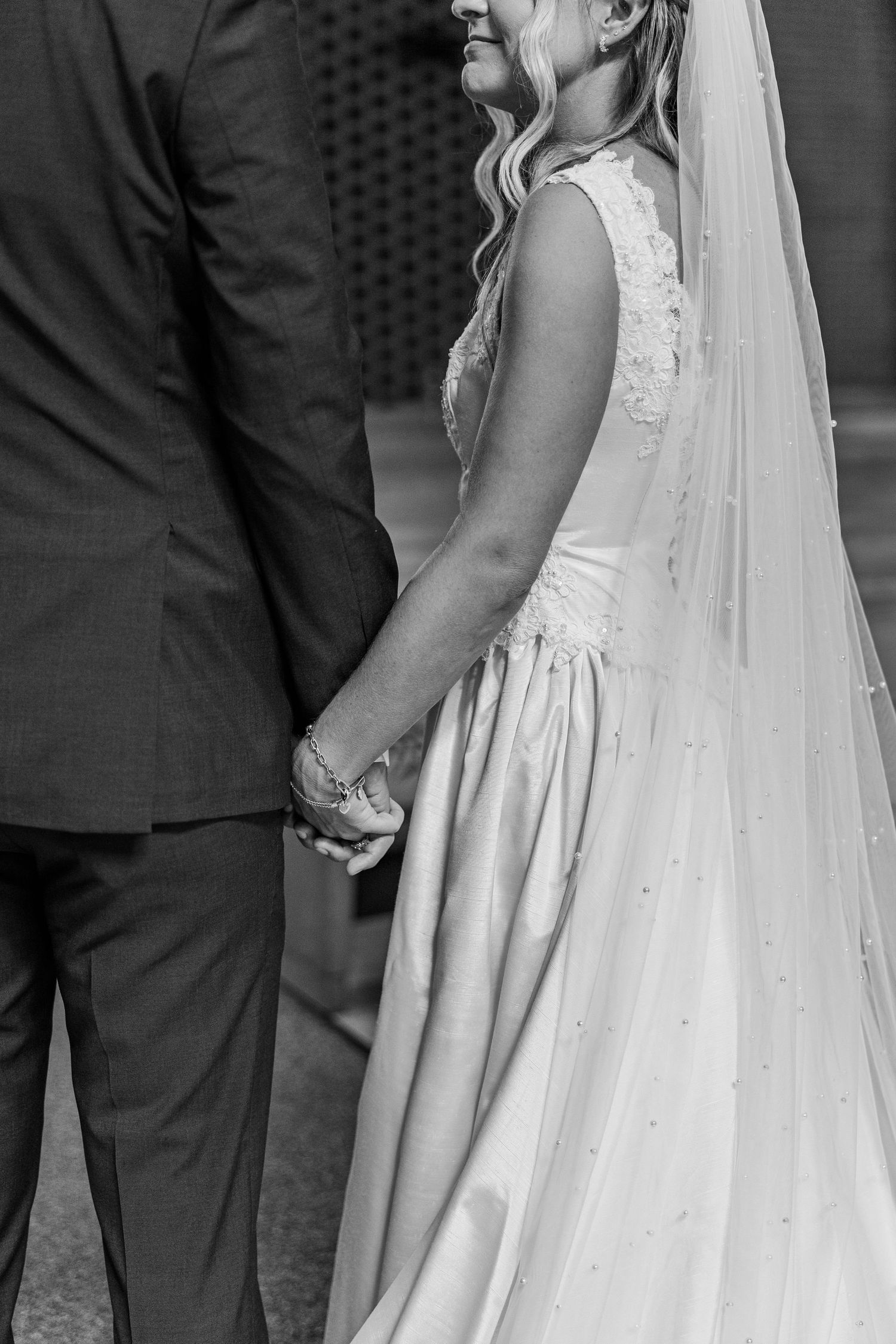 Black and white photo of bride and groom holding hands, bride wearing lace wedding gown with veil.