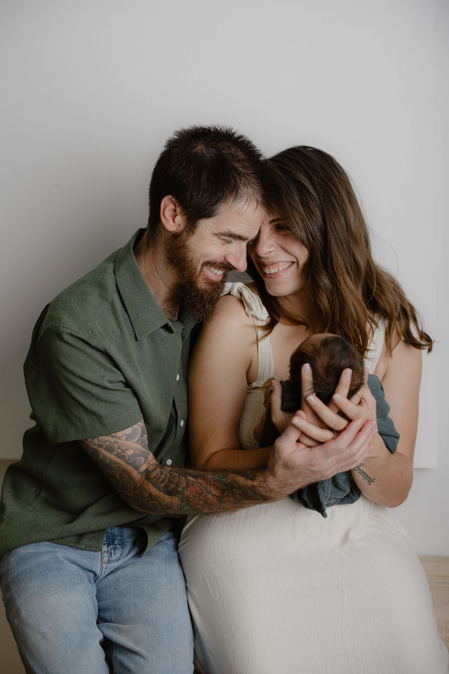 Happy couple smiling and cuddling their newborn baby, man with tattoos in green shirt, woman in white dress.