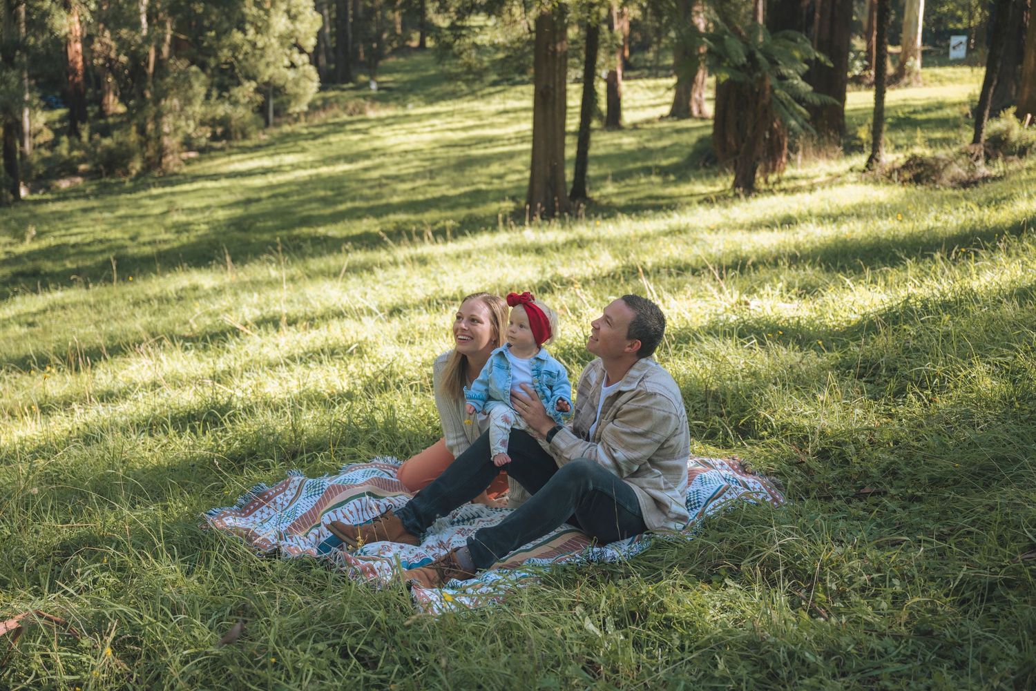 Family enjoying a sunny picnic on a blanket in a lush green forest park setting.