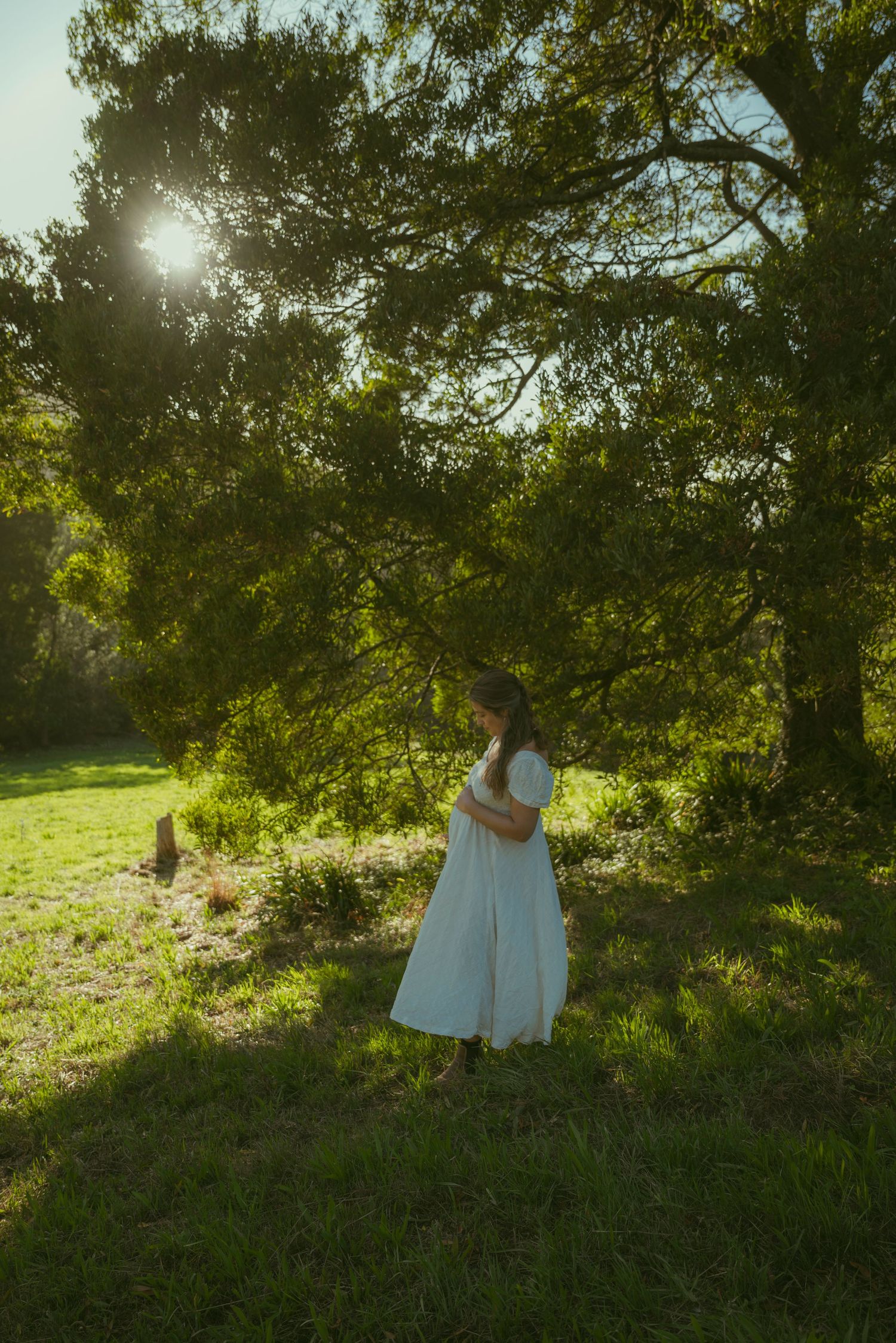 Woman in white dress standing under sunlit green trees in a lush garden setting.