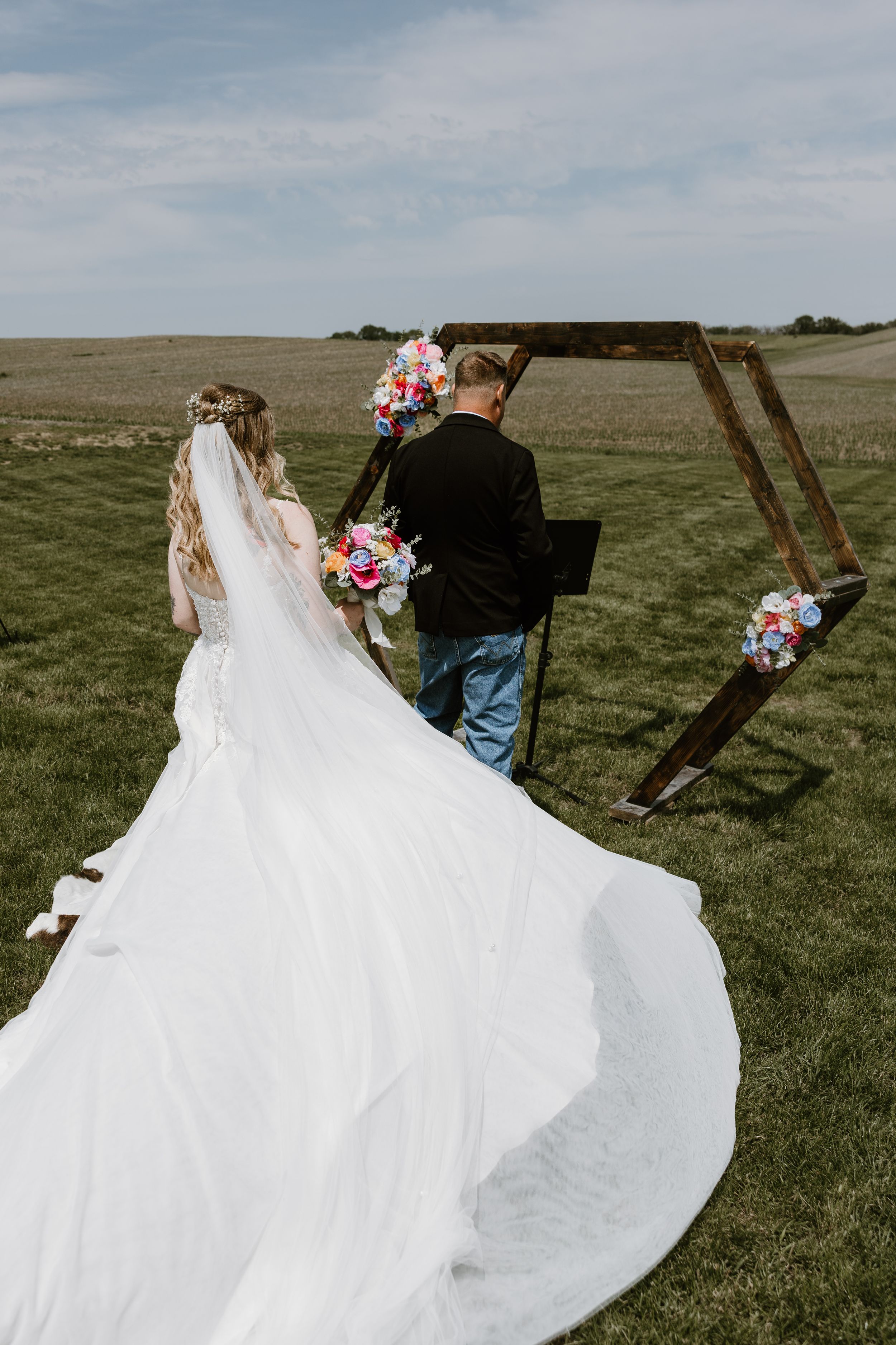 Bride and groom walking toward a rustic wooden arch in an open field, bride holding colorful bouquet.