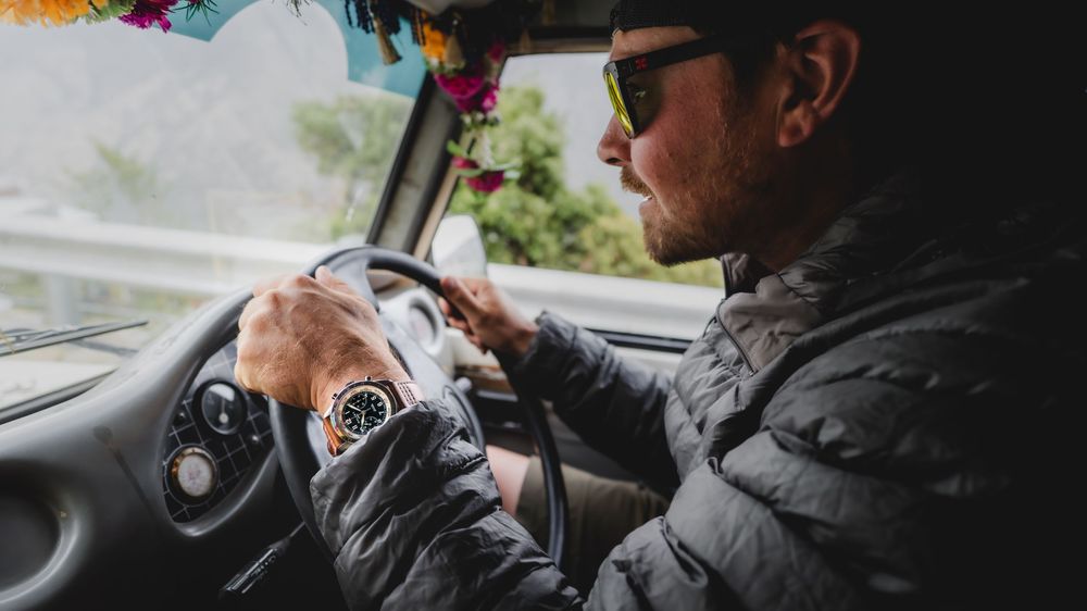 Man wearing sunglasses and a watch driving a vehicle, gripping the steering wheel on a scenic road trip.