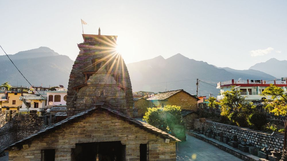 Ancient stone temple tower with sun rays at golden hour, mountains and colorful village buildings in background.