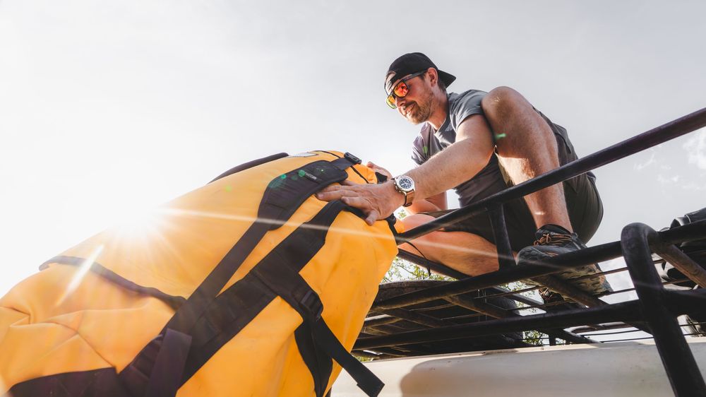 Man securing a yellow duffel bag on a roof rack in bright sunlight, wearing sunglasses and a watch.