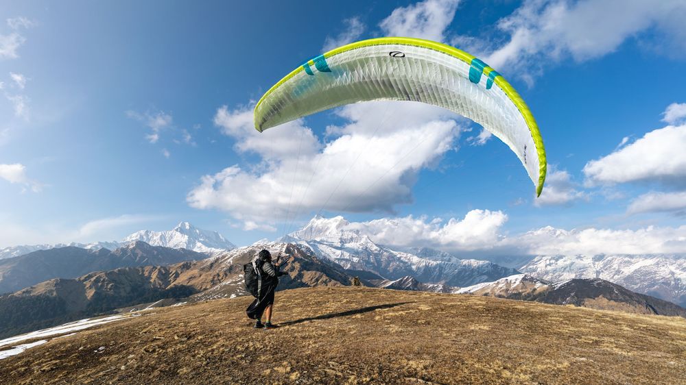 Paraglider launching from a mountain summit with snow-capped peaks and blue sky in the background.