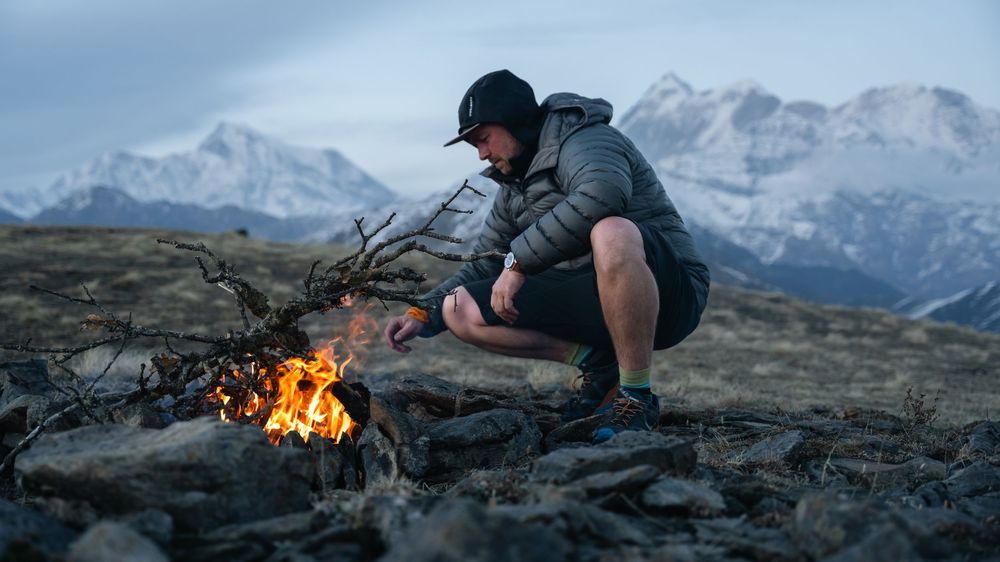 Man in puffer jacket crouching by small campfire with snow-capped mountain peaks in background at dusk.