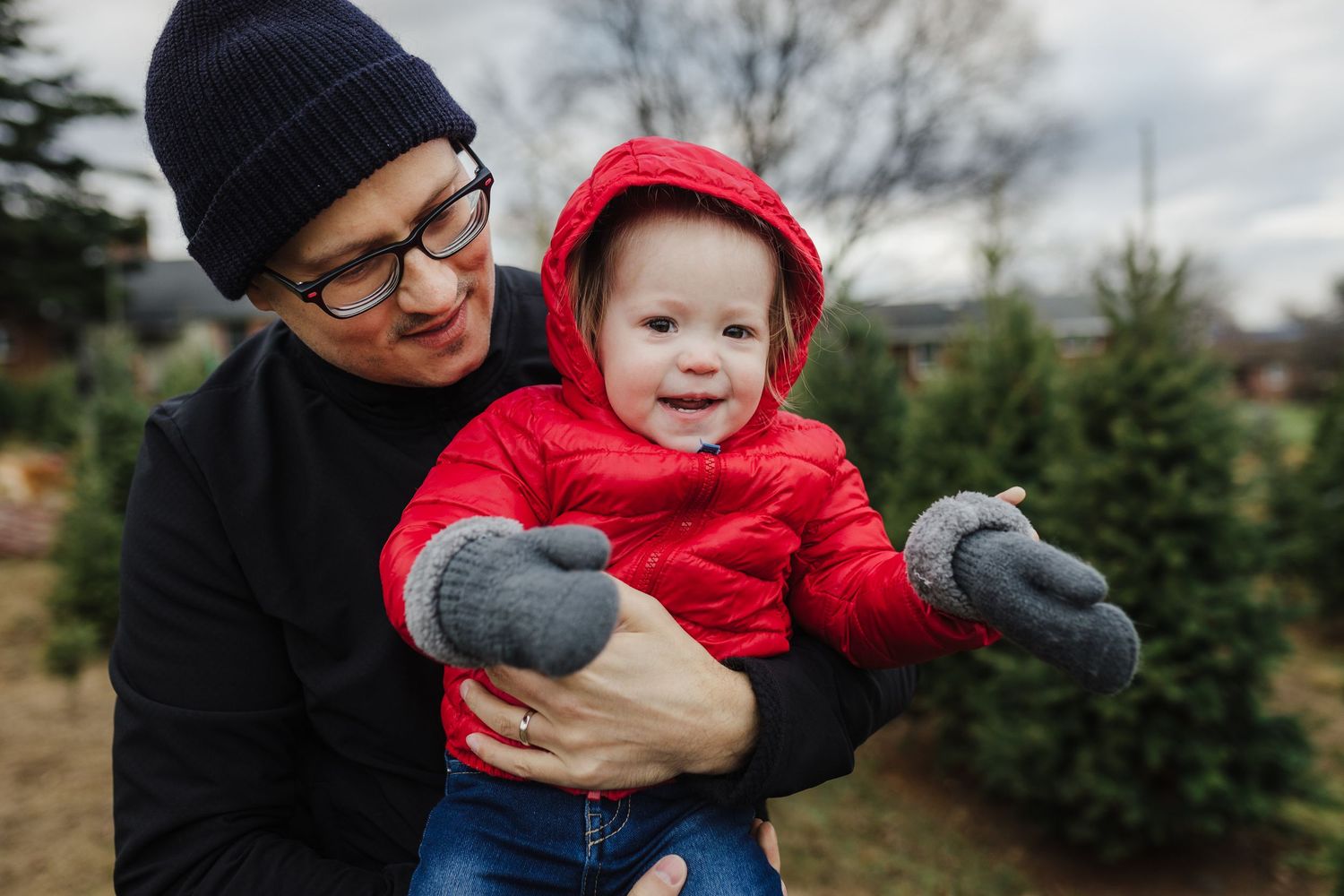 A parent holding a smiling baby in a red hooded jacket at a Christmas tree farm.