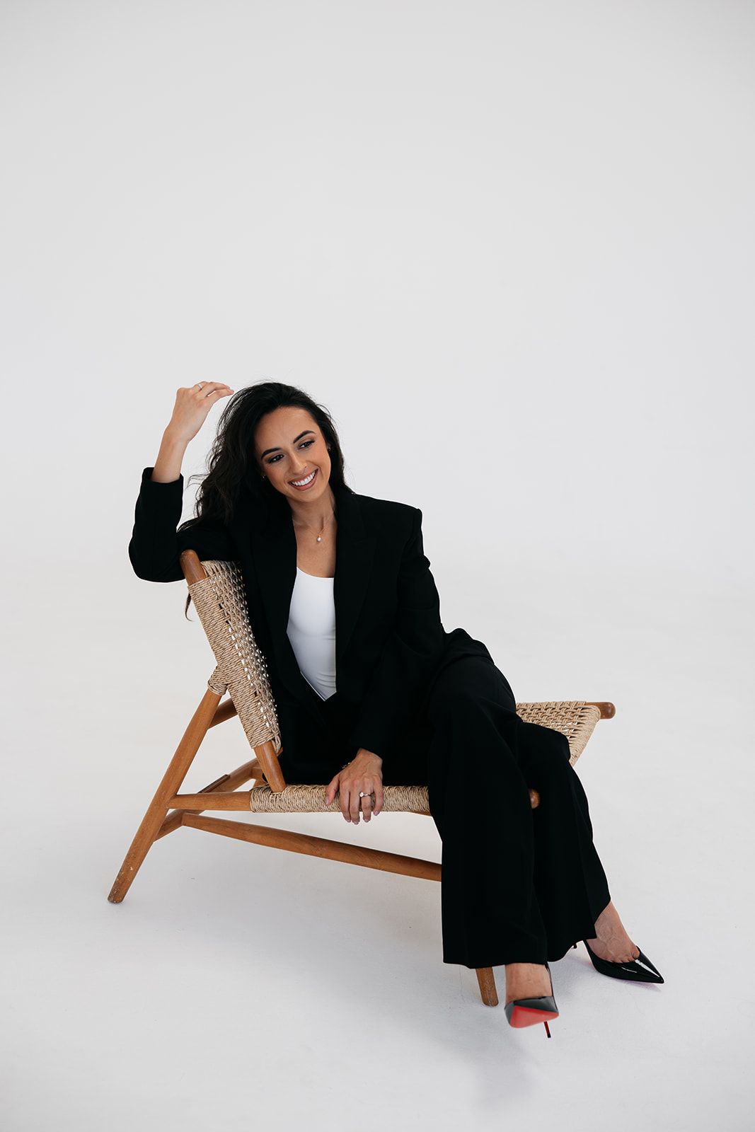Woman in black suit sitting relaxed on woven chair, smiling confidently against white background.