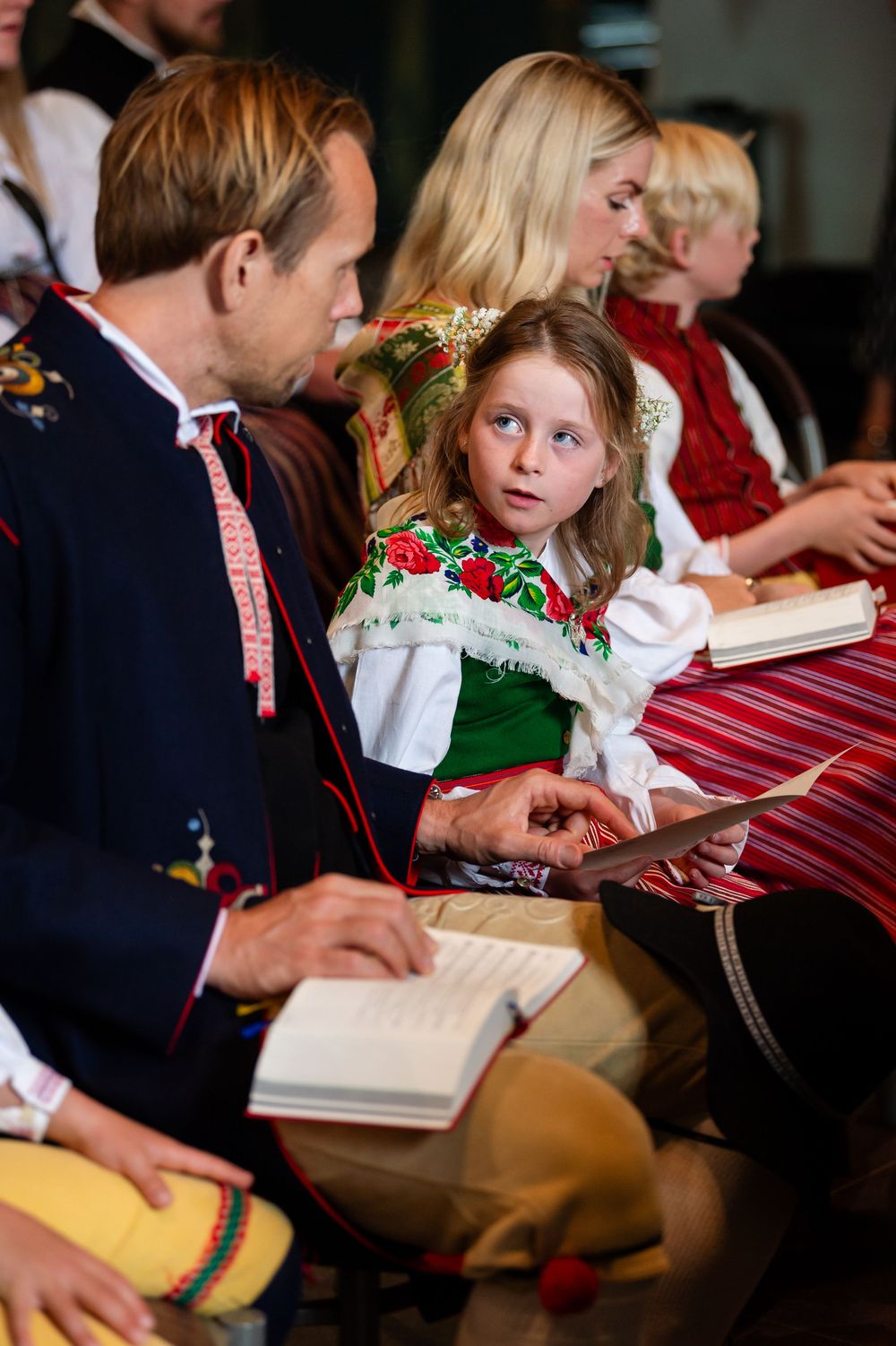 Children and adults in traditional Scandinavian folk costumes sit together reading books at a cultural celebration.