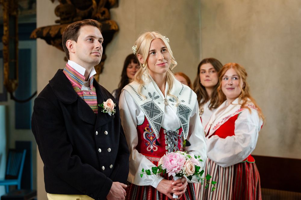 Couple in traditional Scandinavian folk costumes at a wedding ceremony, with guests in matching attire in the background.