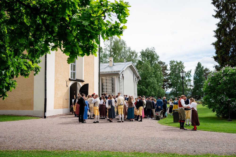 Group of people in traditional folk costumes gathered outside a yellow historic building surrounded by lush green trees.