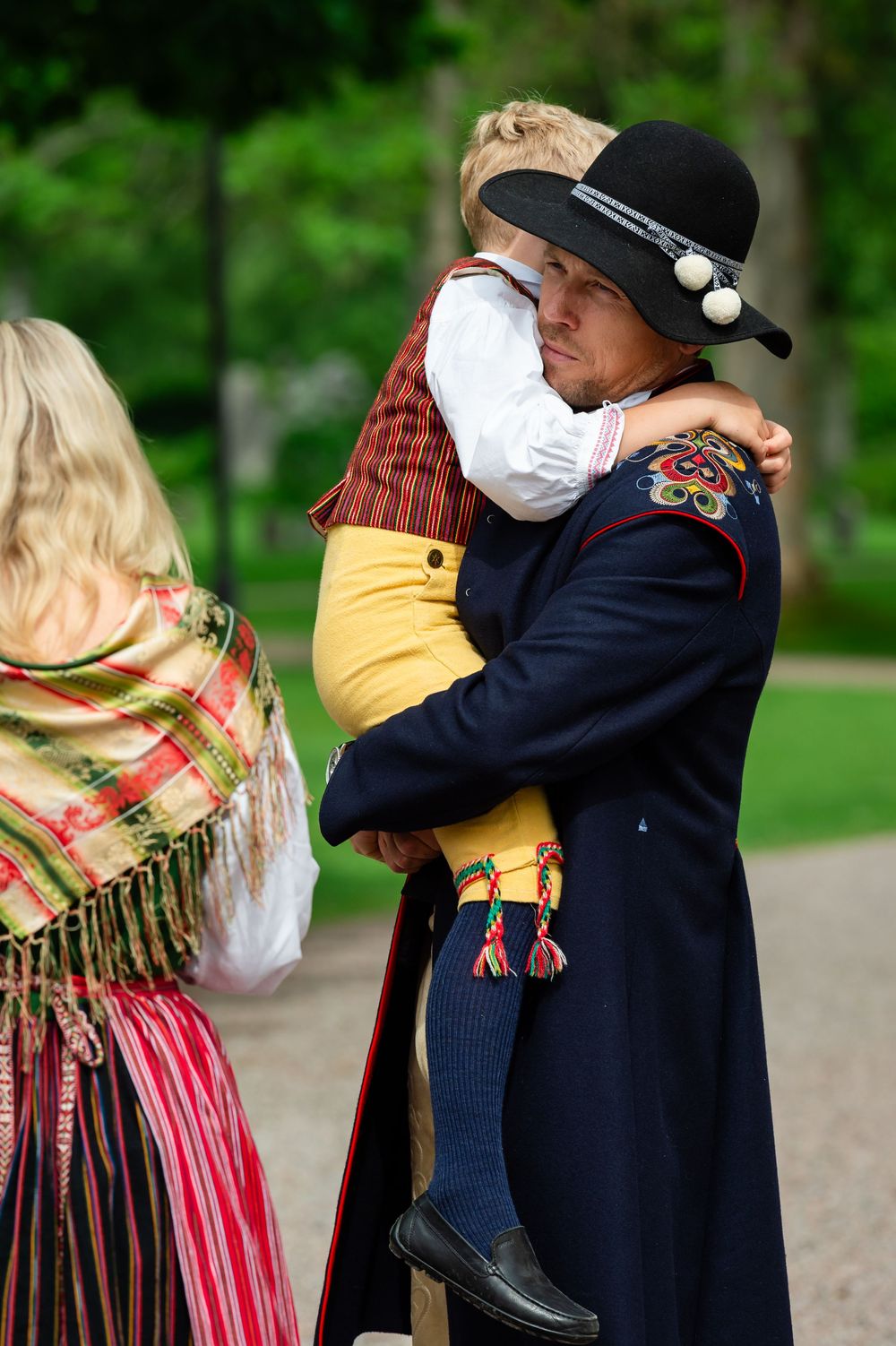 Man in traditional Scandinavian folk costume and black hat tenderly holds a child in matching traditional attire outdoors.