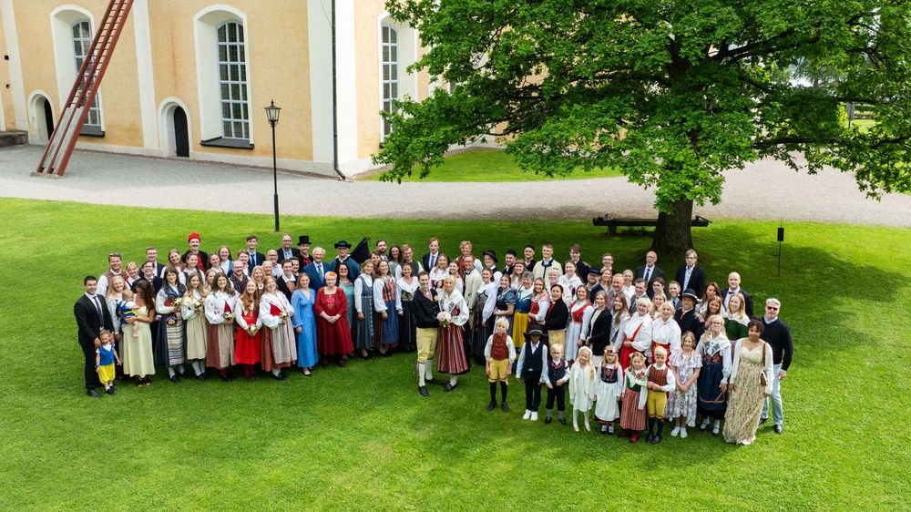 Large group of people in traditional folk costumes gathered on green lawn outside historic white building.
