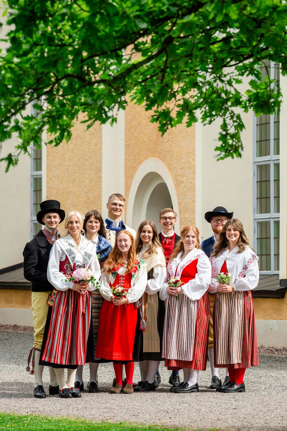 Group of people in traditional Nordic folk costumes posing outdoors near a historic building on a sunny day.