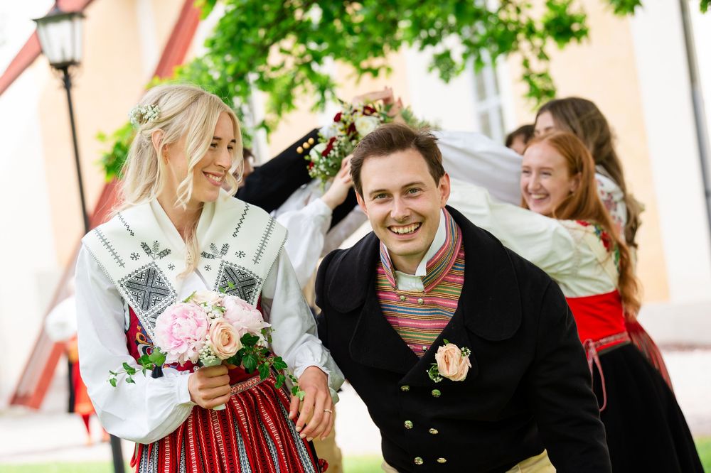 Couple in traditional Nordic folk costumes smiling outdoors, bride holding pink bouquet, guests in festive attire in background.