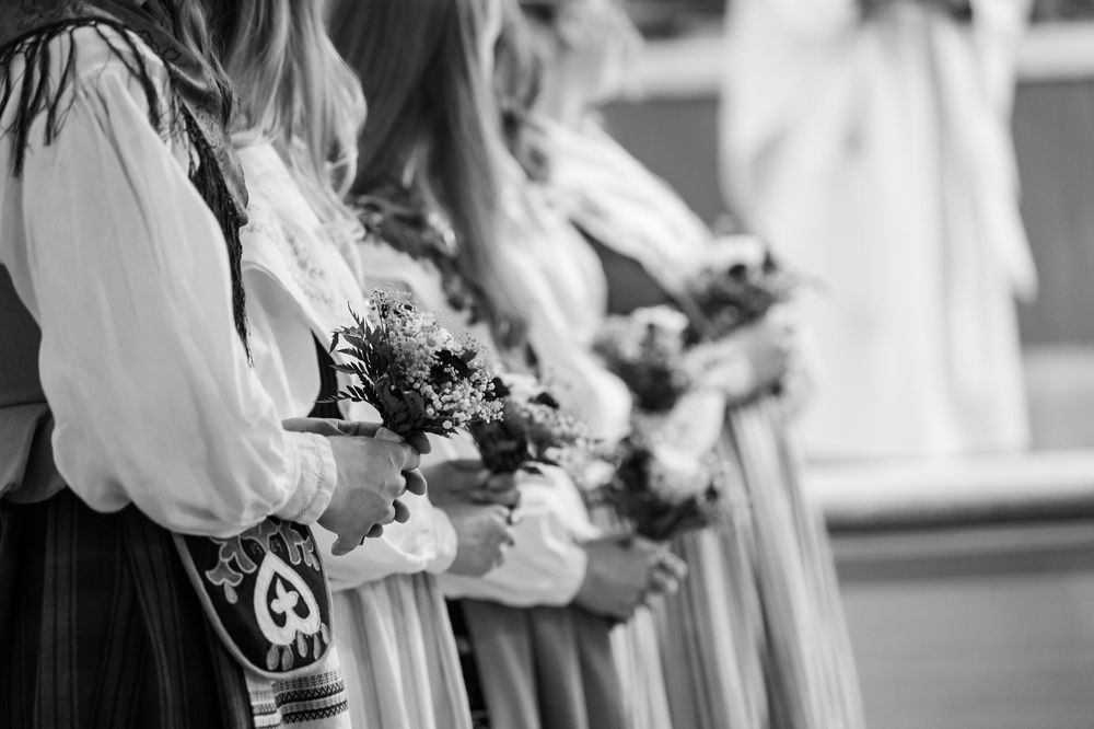 Women in traditional folk costumes holding bouquets of flowers in a black and white ceremonial photo.