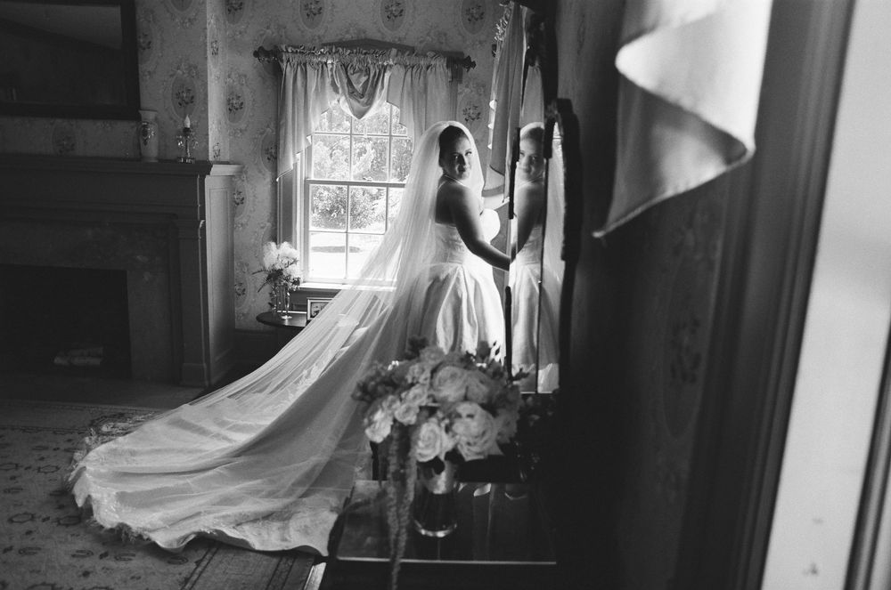 Black and white photo of a bride in a flowing gown and veil standing by a window with bouquet nearby.