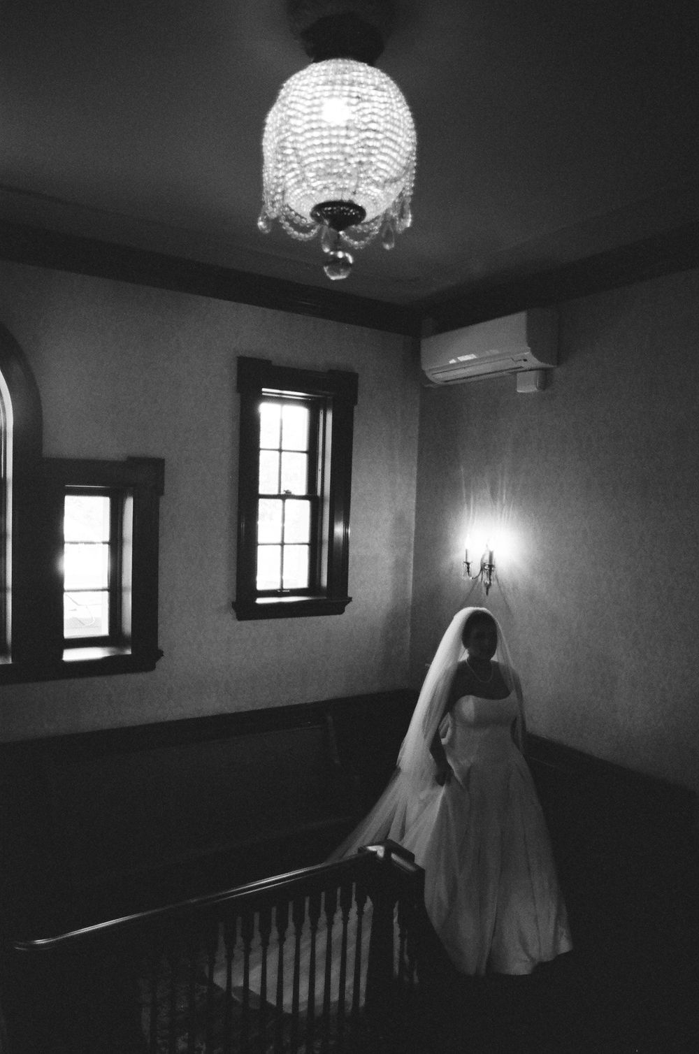 Bride in white wedding gown and veil stands on staircase beneath crystal chandelier in black and white photo.