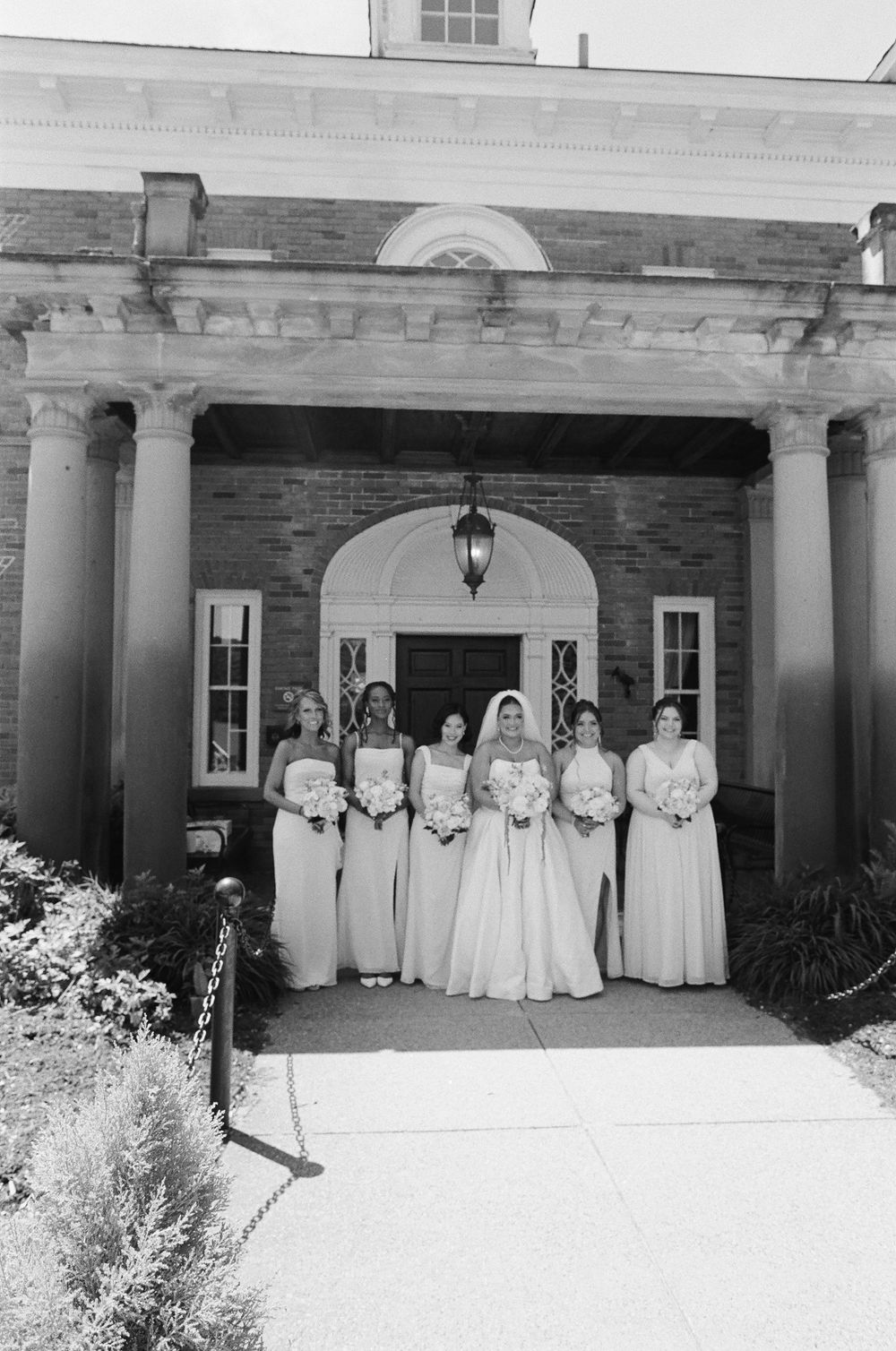 Bride and bridesmaids in white gowns pose before a brick colonial building with columns in a black and white photo.