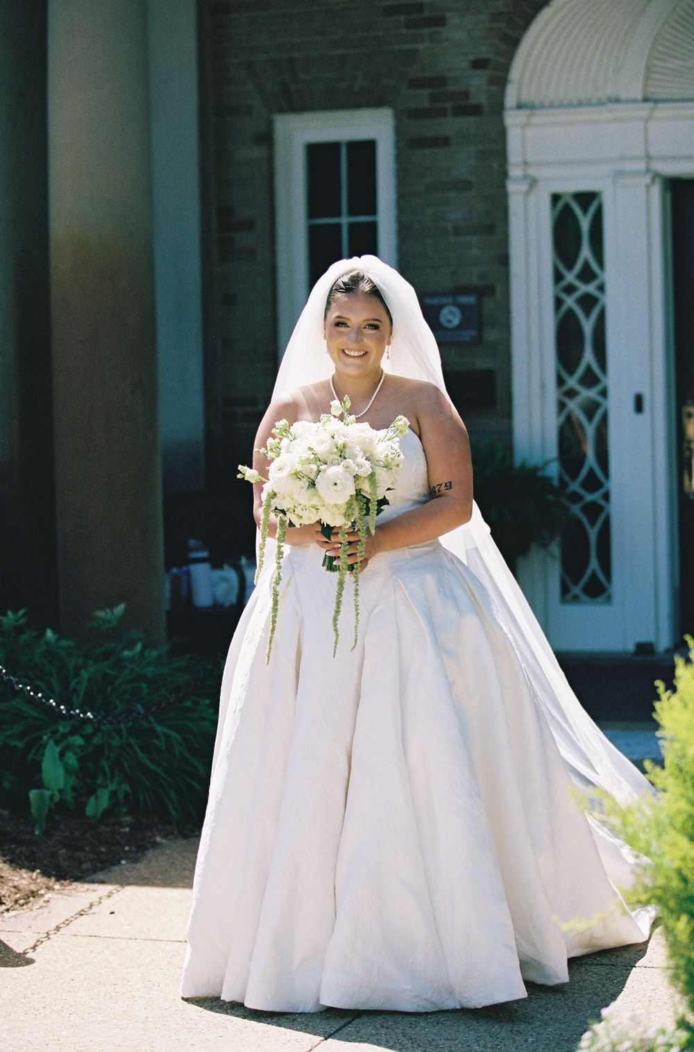 Smiling bride in strapless ball gown holding white bouquet standing outside brick church entrance.