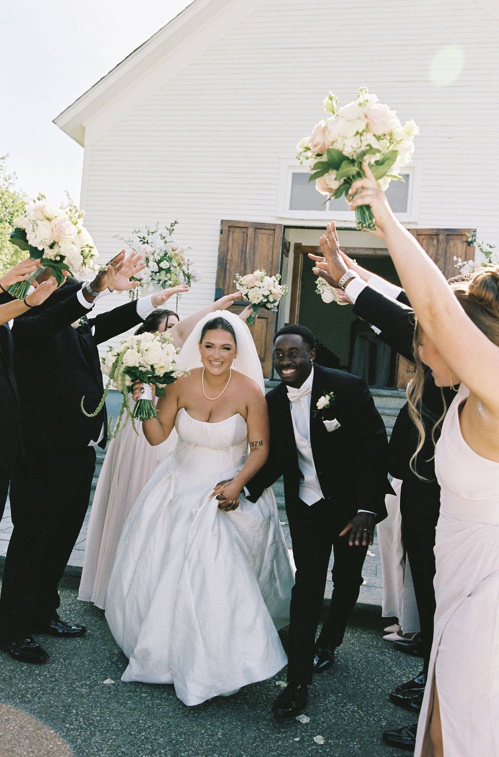 Joyful newlywed couple exits white chapel as wedding party raises bouquets in celebration.