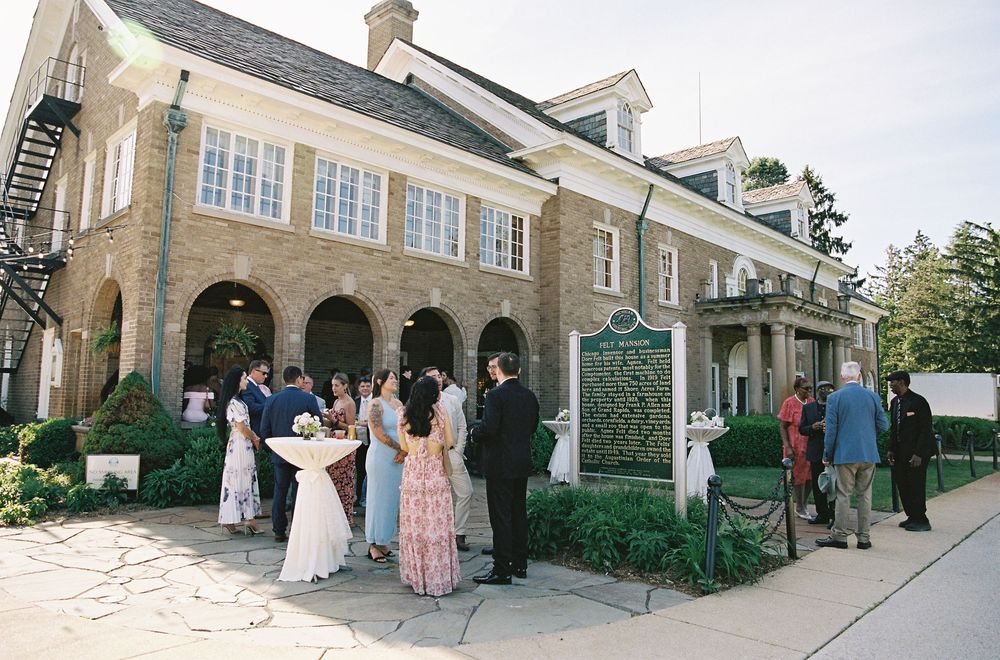 Wedding guests mingle outside elegant stone manor house venue during outdoor cocktail reception.