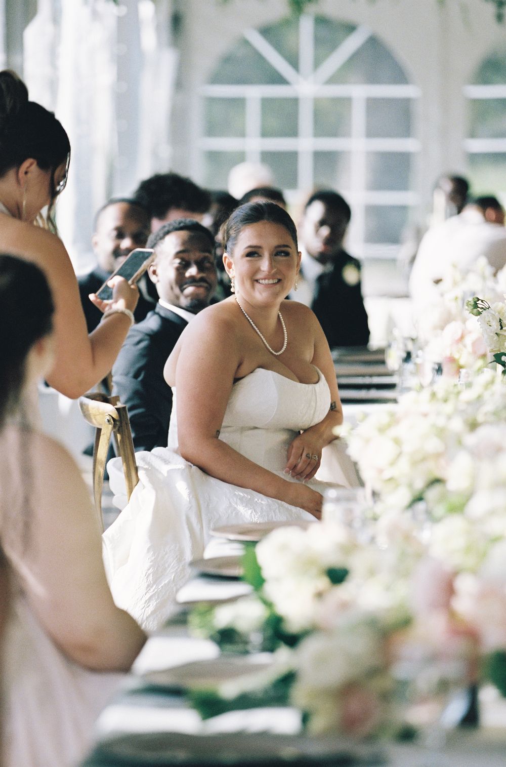 Smiling bride in strapless gown seated at elegant floral-adorned reception table with guests nearby.
