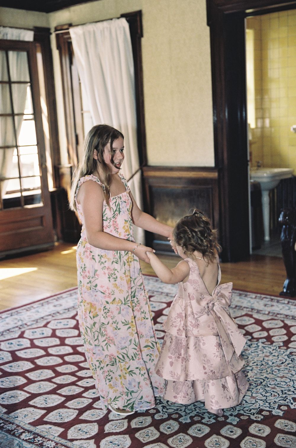 Bridesmaid in floral maxi dress dances with young flower girl in pink dress indoors.