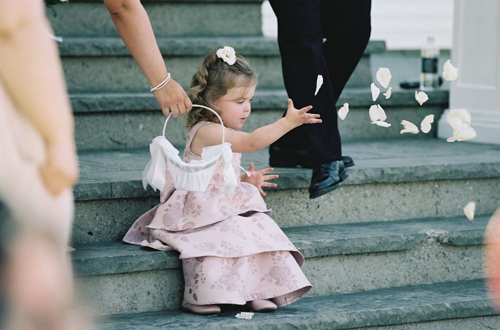 Young flower girl in pink dress tosses petals on stone steps during wedding ceremony.