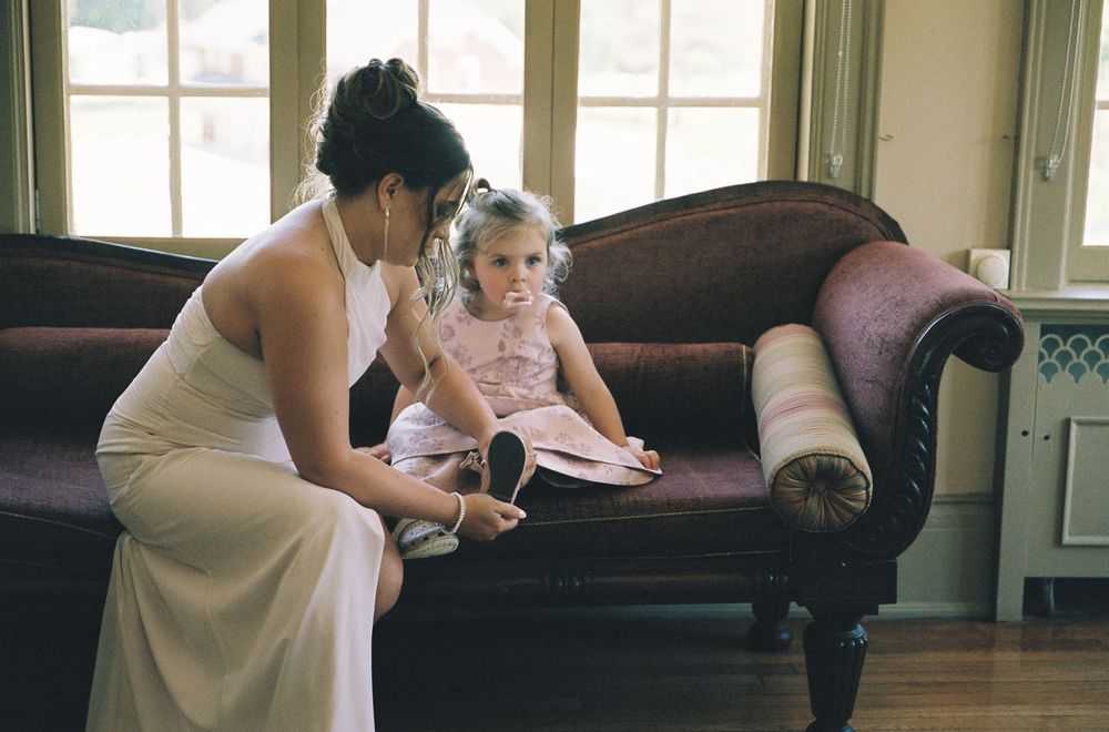 Bridesmaid in white dress helping young flower girl with shoes on vintage velvet sofa.