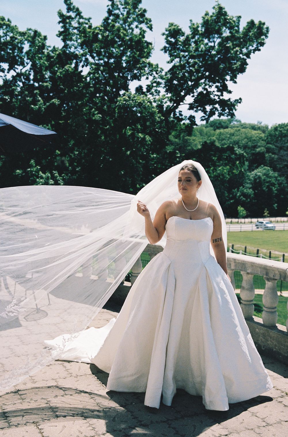 Bride in a strapless satin ball gown holding her flowing veil on a stone terrace outdoors.