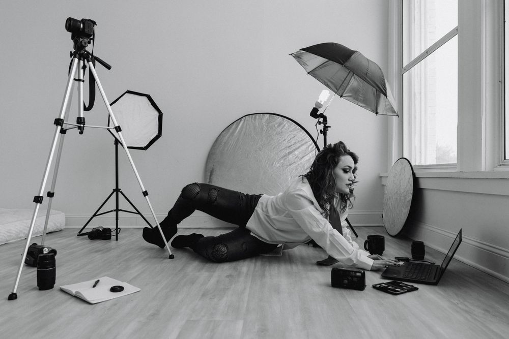 Woman wearing a button down shirt & tie, sitting on the floor using a laptop surrounded by photography gear