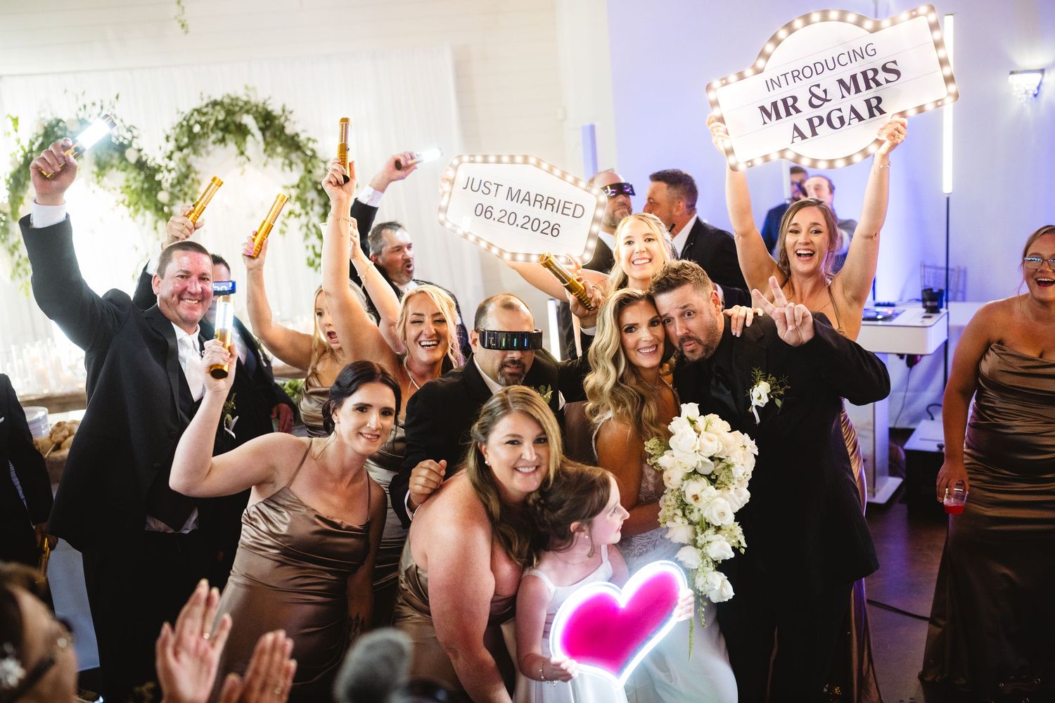 Joyful wedding reception group photo with guests celebrating, holding Mr. & Mrs. Apgar signs and Just Married banner.