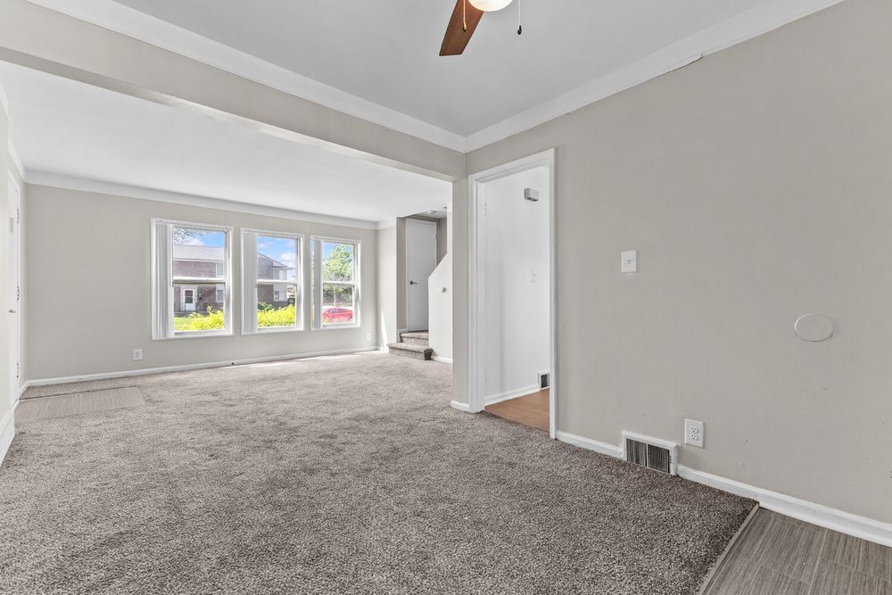 Spacious living room with gray carpet, crown molding, ceiling fan, and large windows offering natural light.