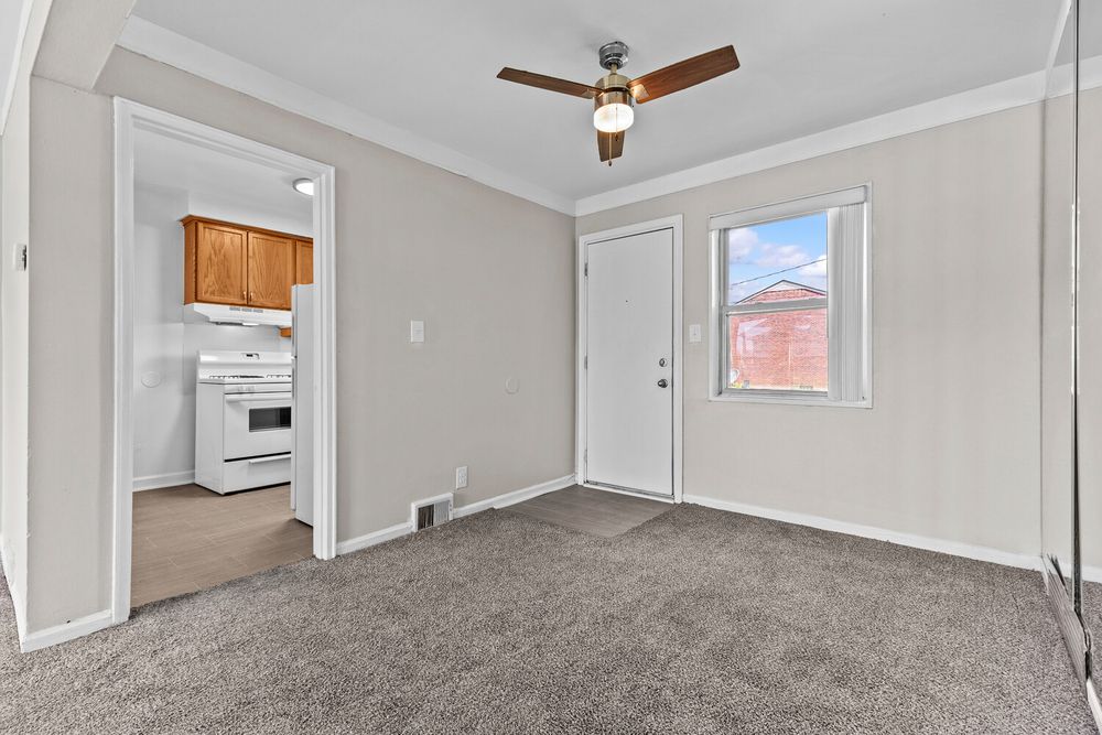 Bright living room with gray carpet, ceiling fan, crown molding, and view into kitchen with white stove.