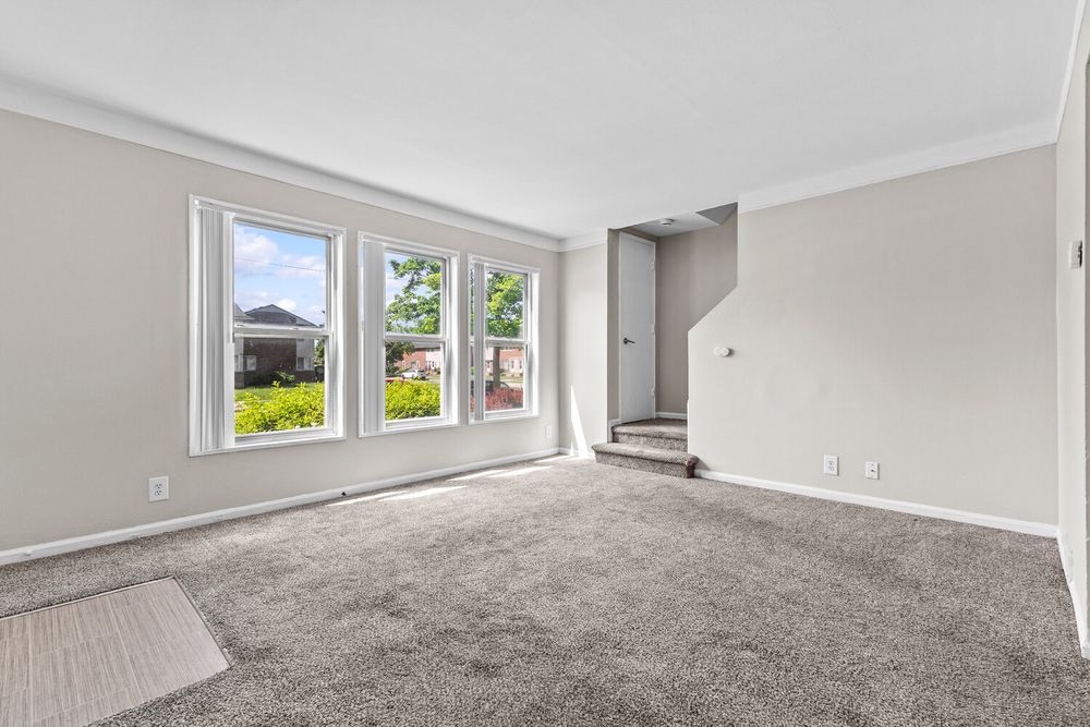Bright empty living room featuring plush gray carpet, crown molding, and large windows with garden views.