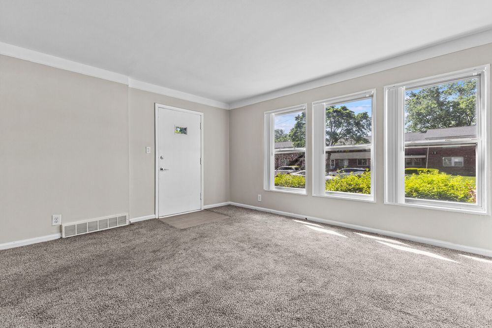 Bright living room with large windows, gray carpet, white walls, crown molding, and a view of lush greenery outside.