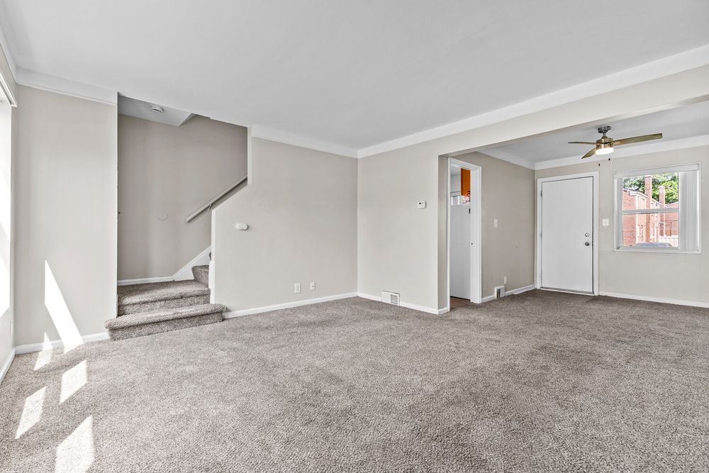 Open living area with gray carpet, crown molding, ceiling fan, and staircase leading to upper level.