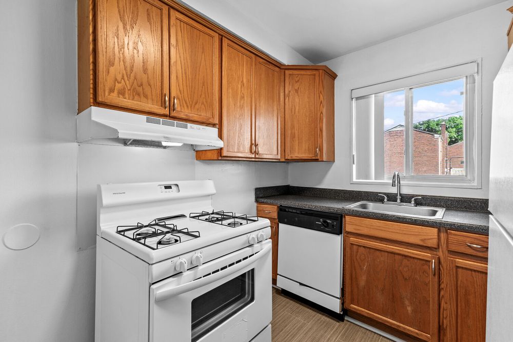 Kitchen with wood cabinets, white gas range, white dishwasher, dark countertops, and a window above the sink.