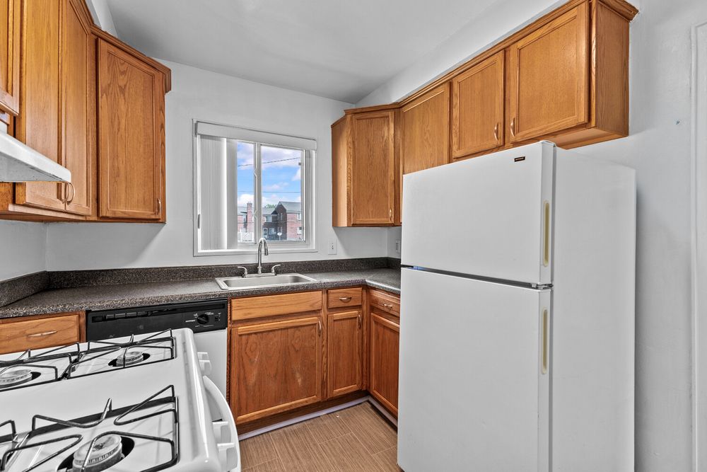 Compact kitchen with wood cabinets, white refrigerator, gas stove, dark countertops, and a window above the sink.