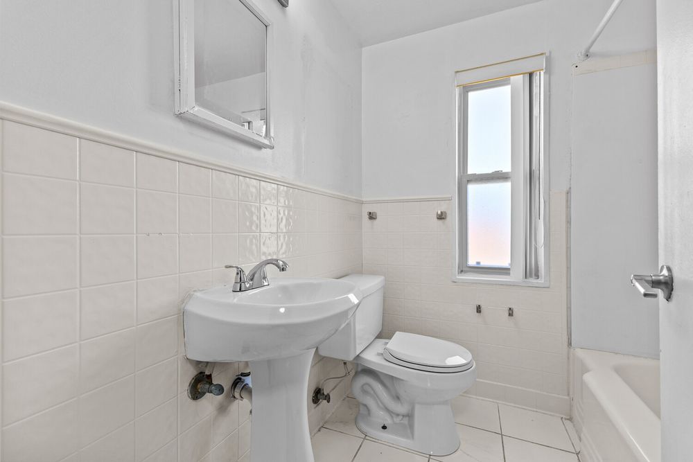 Clean white bathroom with pedestal sink, toilet, tiled walls, and bathtub next to a bright window.
