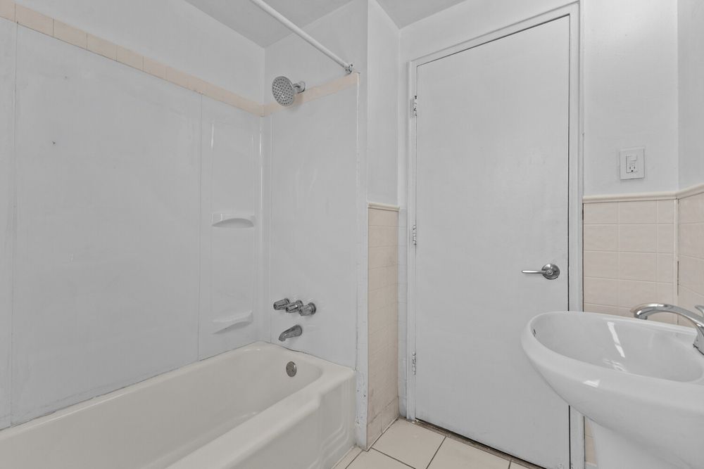 Simple white bathroom with bathtub, shower, pedestal sink, and white door on light tile flooring.