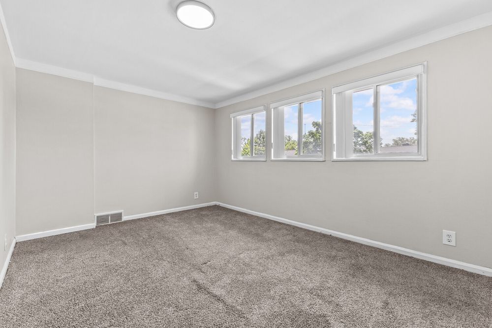 Bright empty bedroom with gray carpet, crown molding, and three windows with natural light.